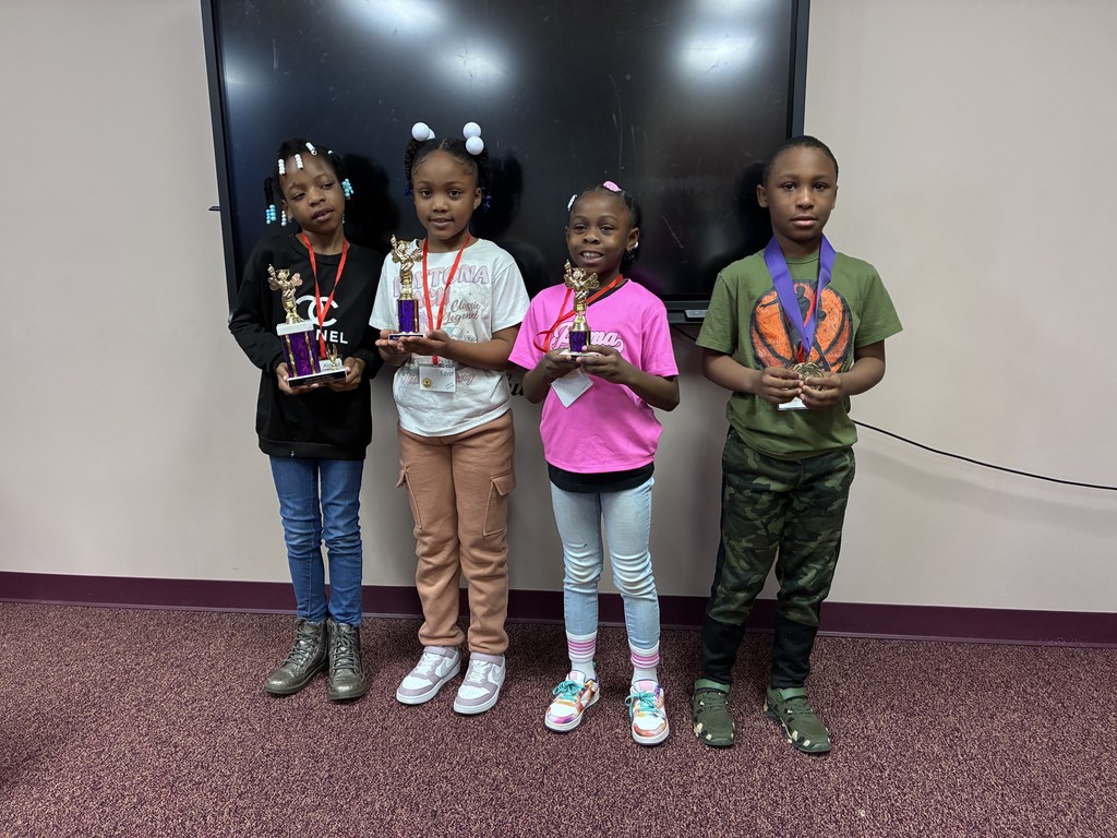 Four children in a room, each holding a trophy, smiling, and standing close to each other.
