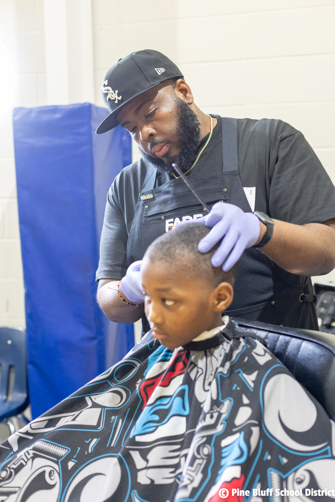 A man cuts a child's hair in a classroom setting. The child is seated, wearing a colorful cape.