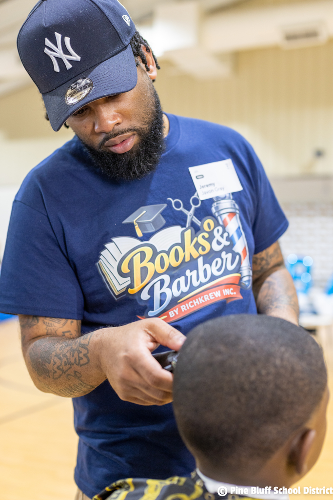 A man with a cap trims a boy's hair. The man's shirt reads "Books & Barber".