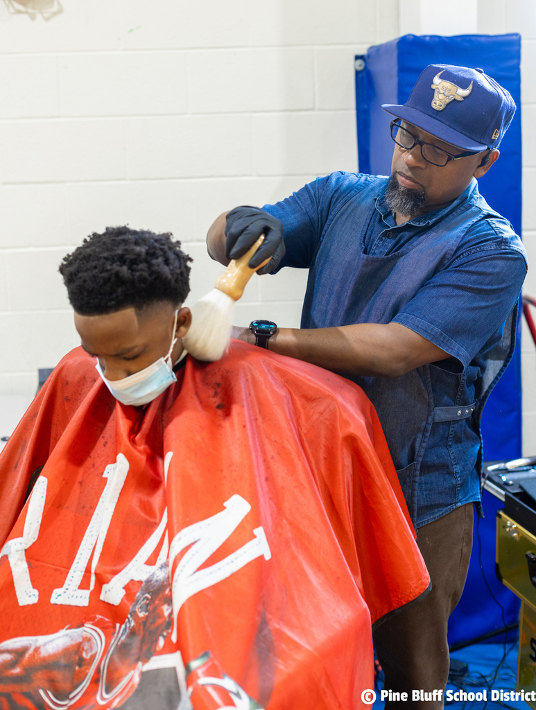 A man in a blue shirt and cap cuts another man's hair. The seated man wears a face mask and an orange cape.