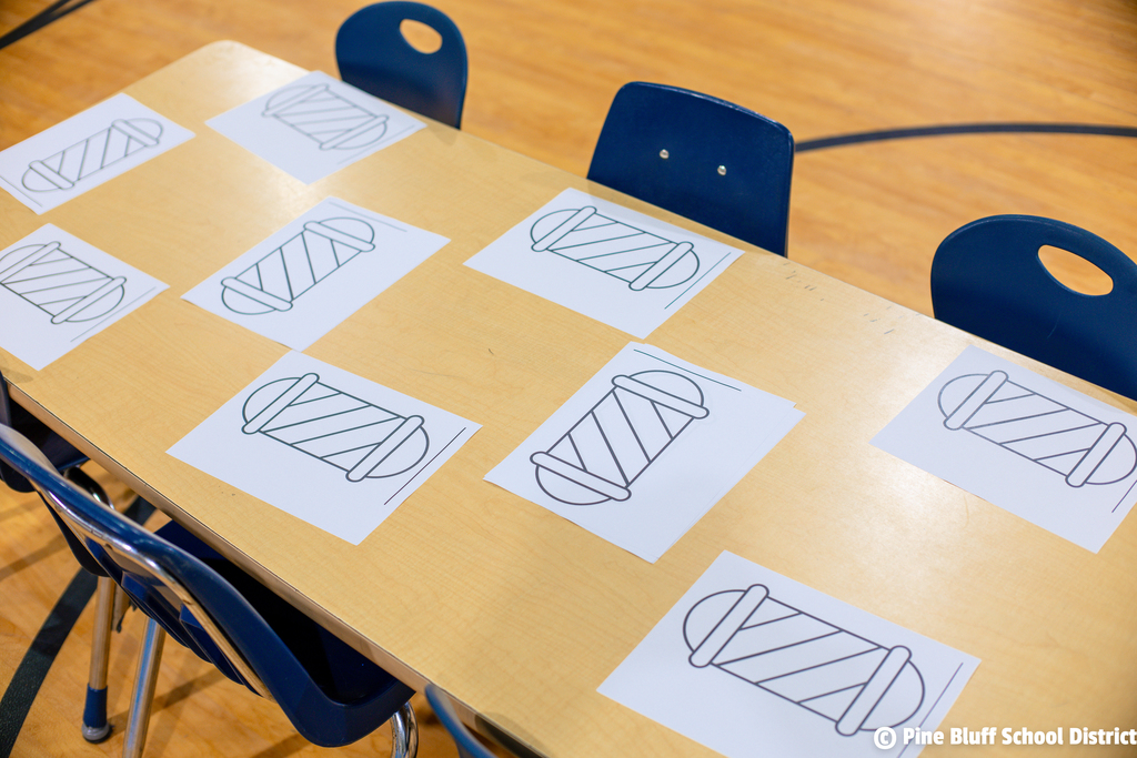 A wooden table holds eight sheets of paper with line drawings. Four blue chairs surround the table. A basketball court floor is in the background.