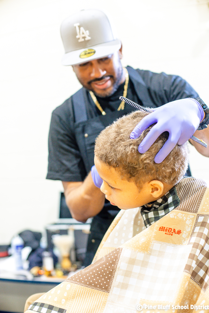A man wearing a baseball cap and gloves cuts a child's hair in a salon.