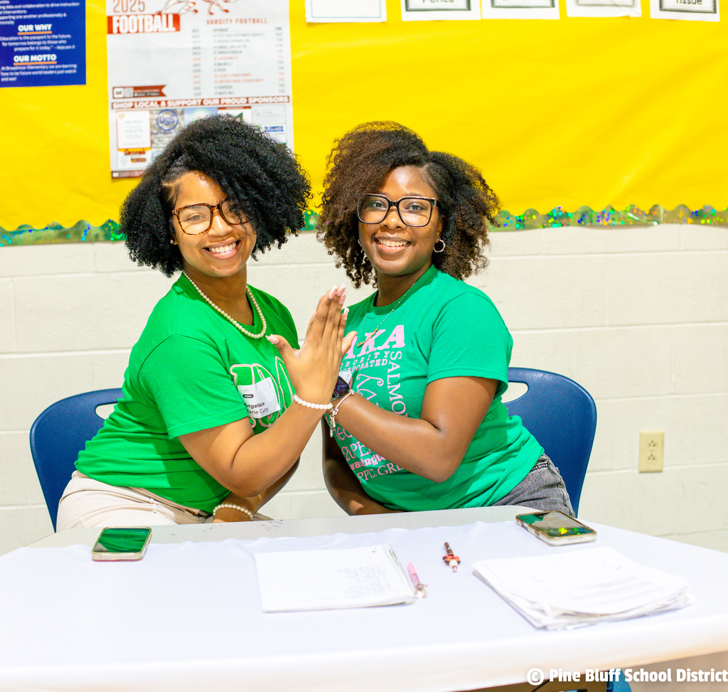 Two women in green shirts sit at a table, smiling, and shaking hands. Papers and cell phones are on the table.