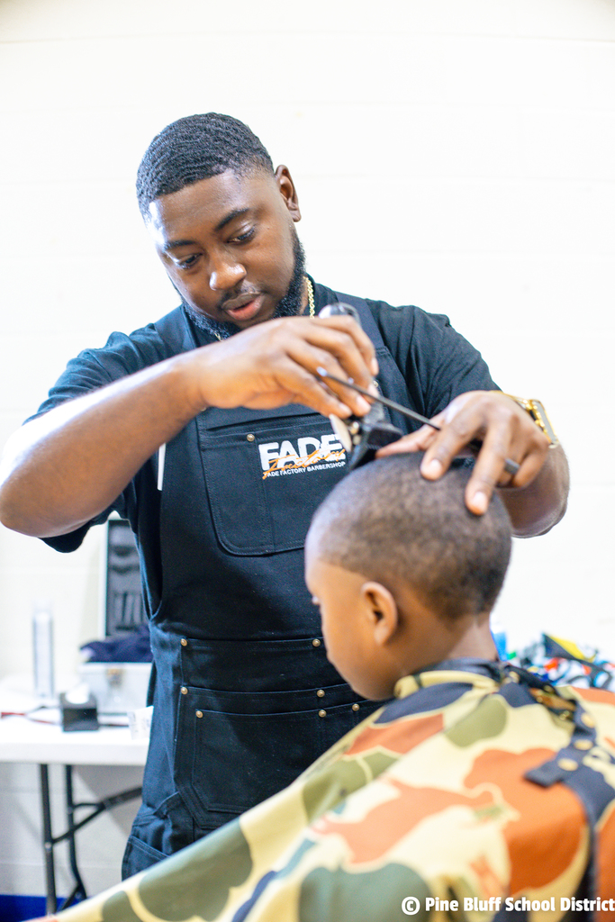 A man with a name tag reading "FADE" cuts the hair of a child in camouflage.
