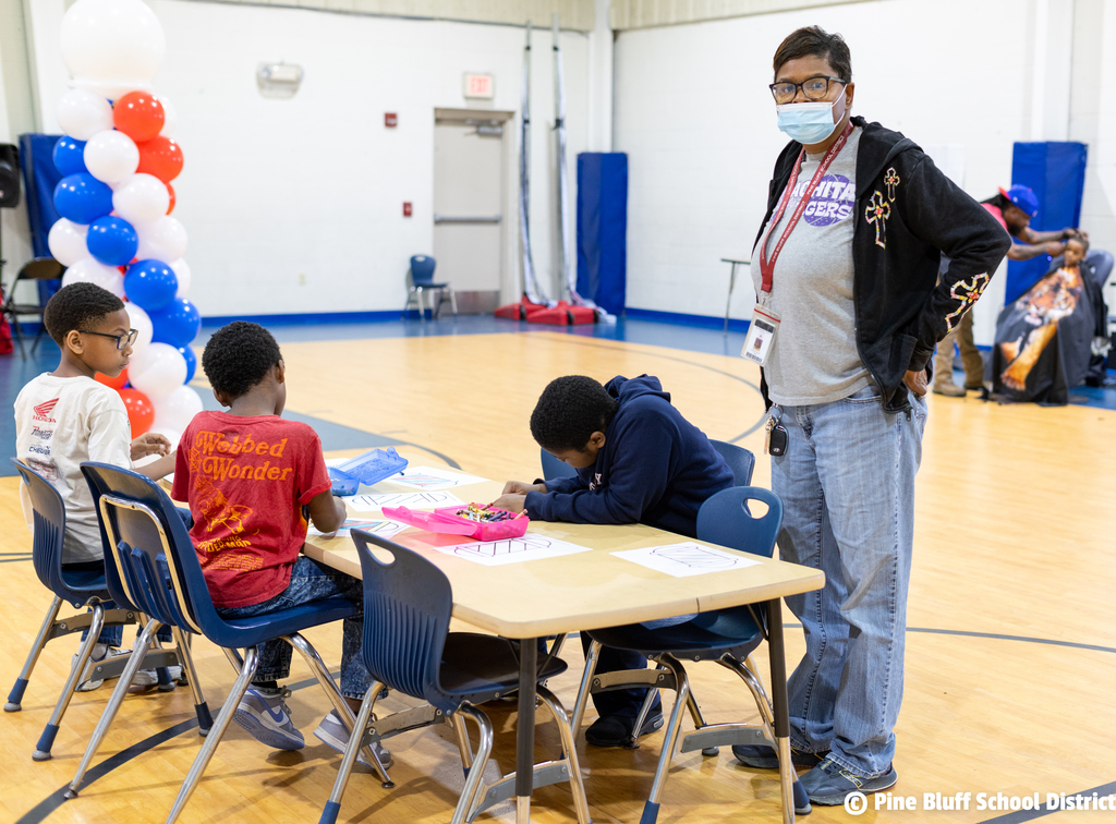 A person in a mask stands at a table in a gym. Three children sit at the table.