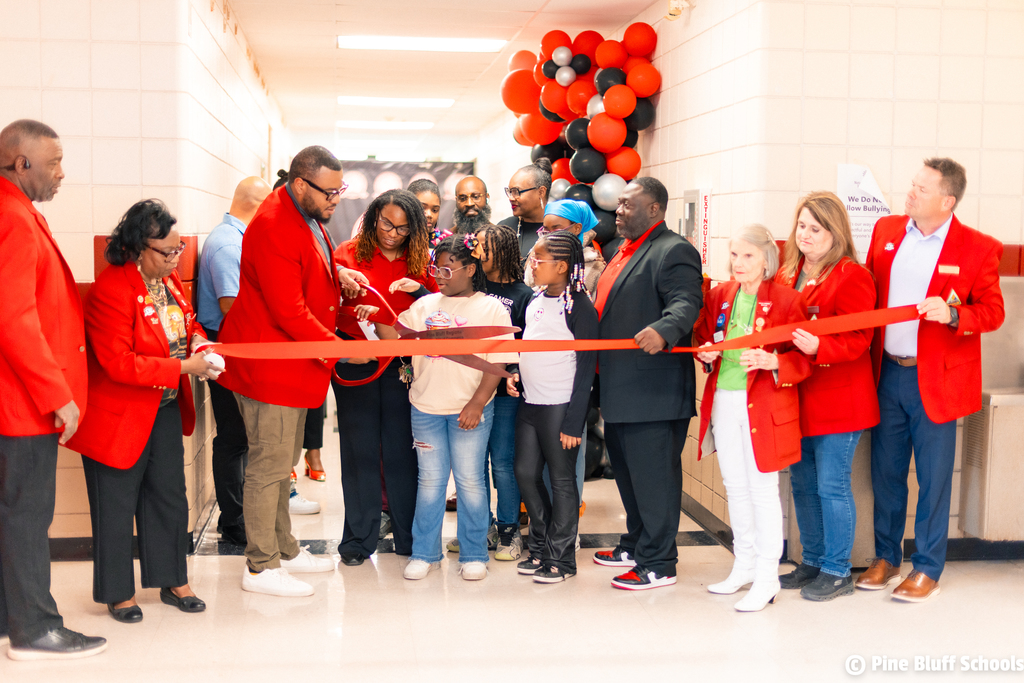 A group of adults and children in red coats stand in a hallway cutting a ribbon.