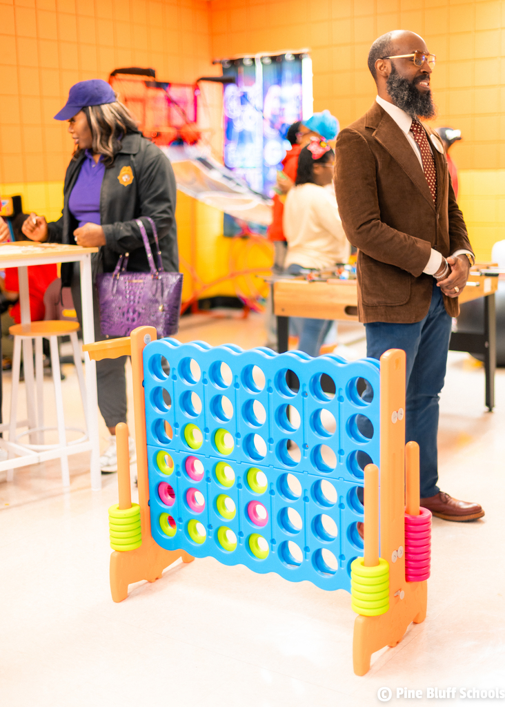 A man and woman in business attire stand near a bright orange wall, facing a table with a Connect Four board.