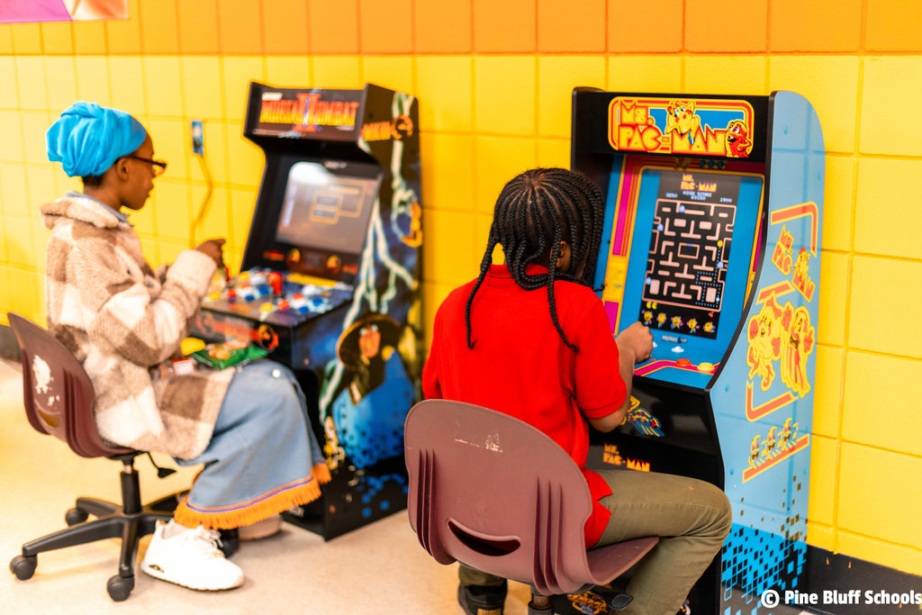 Two individuals play arcade games in a brightly colored room. One wears a blue headscarf, the other a red shirt.
