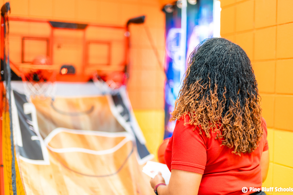 Person with curly hair in red shirt playing basketball game in an indoor facility with an orange wall.