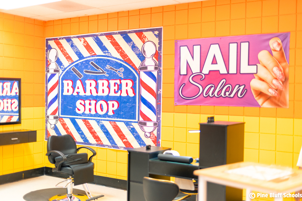 An interior with yellow walls, featuring a barber shop sign and a nail salon banner. Chairs and desks are present.