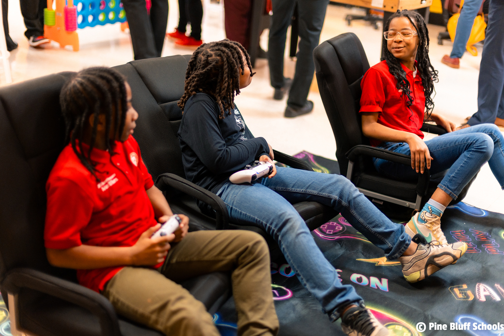 Three students sit in chairs. They wear red shirts and hold game controllers. A rug is on the floor.