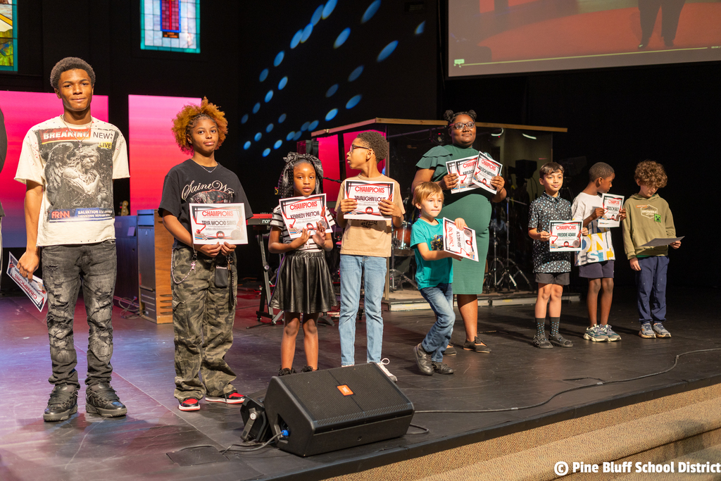 Group of children holding certificates on stage with a speaker and a screen in the background.