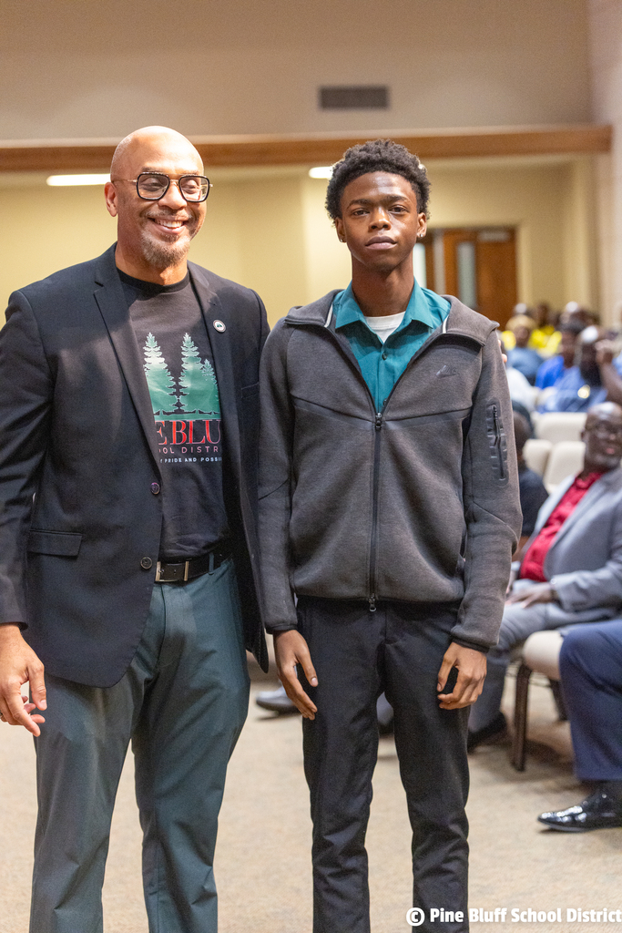 Two men stand together in a room with rows of chairs and people seated behind them.