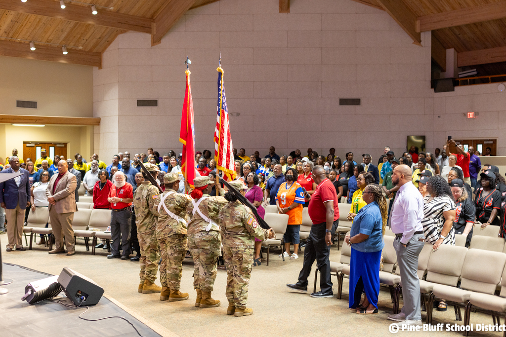 Soldiers standing with flags in a hall. Audience standing in front of them. Speakers on the stage.