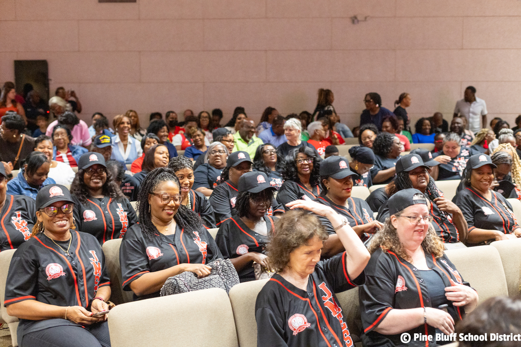 Large group of adults wearing matching red and black uniforms sitting in chairs at an event.