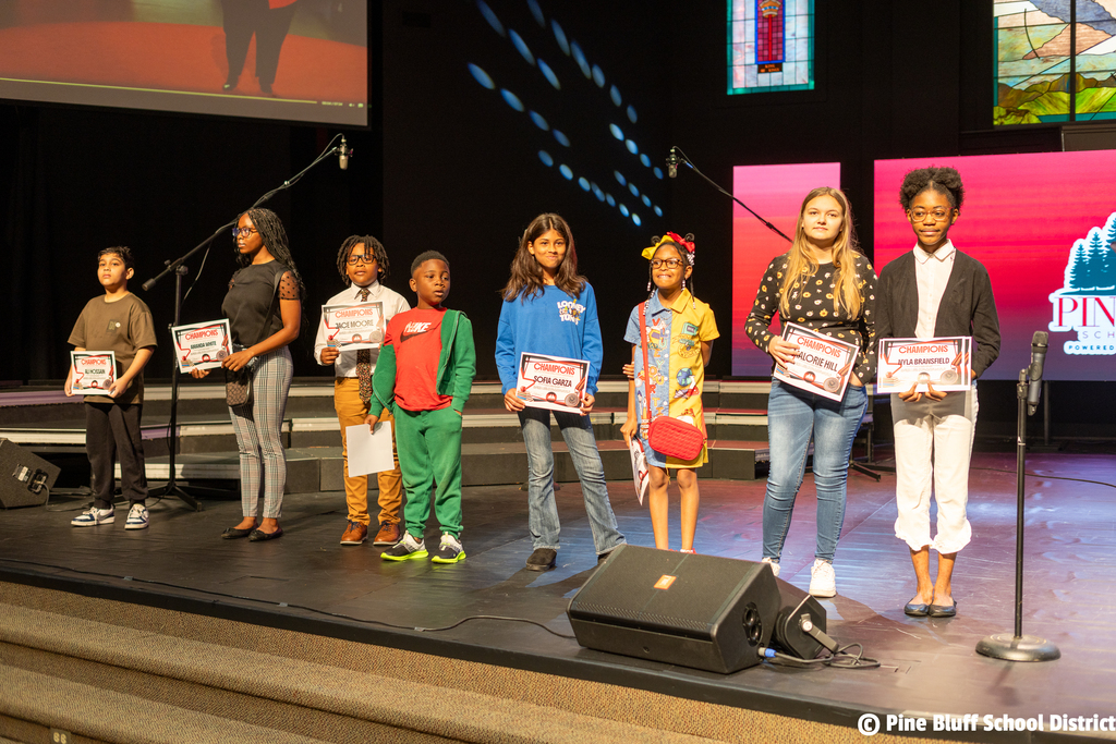 Seven children stand on a stage holding certificates; one has a backpack. Behind them, a microphone and speaker.