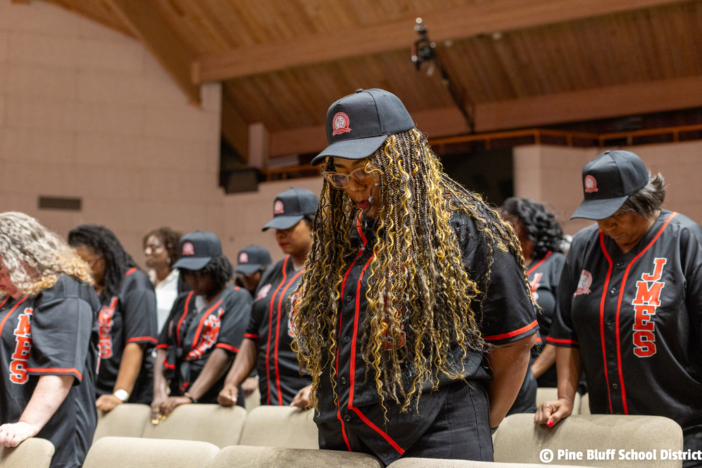 People in black and red baseball jerseys and hats stand in rows, facing front, in a large room.