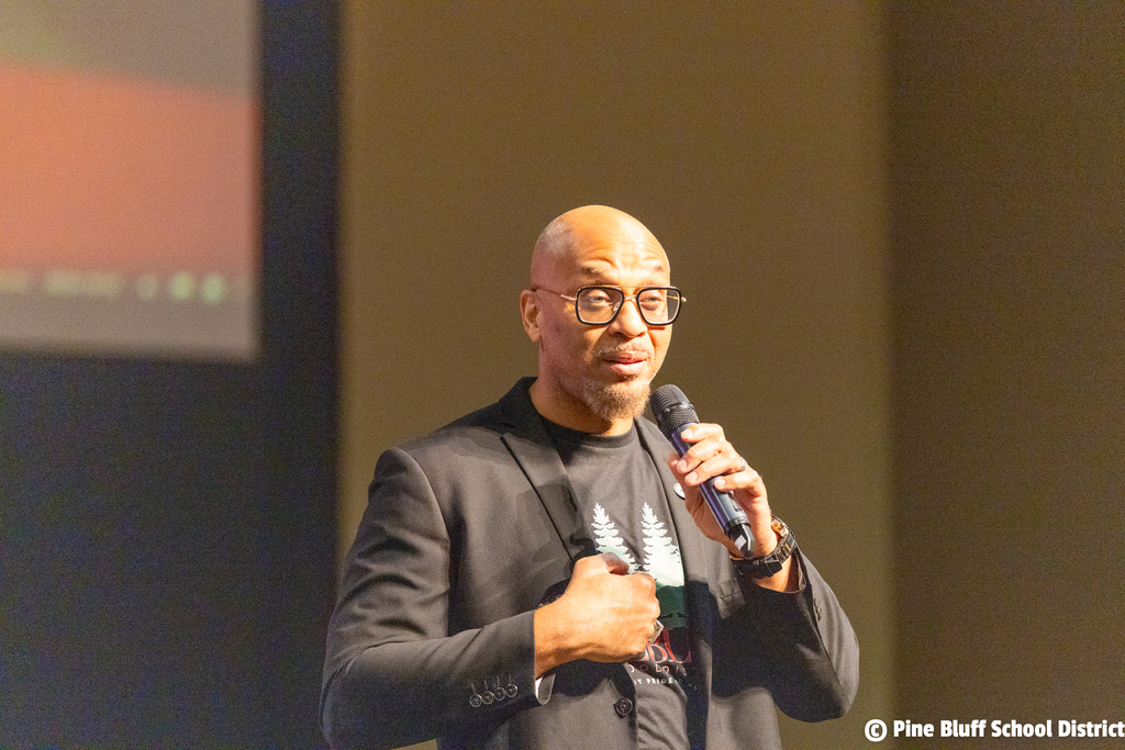 Man speaking into a microphone. He is wearing a dark suit and glasses. Behind him is a large screen.
