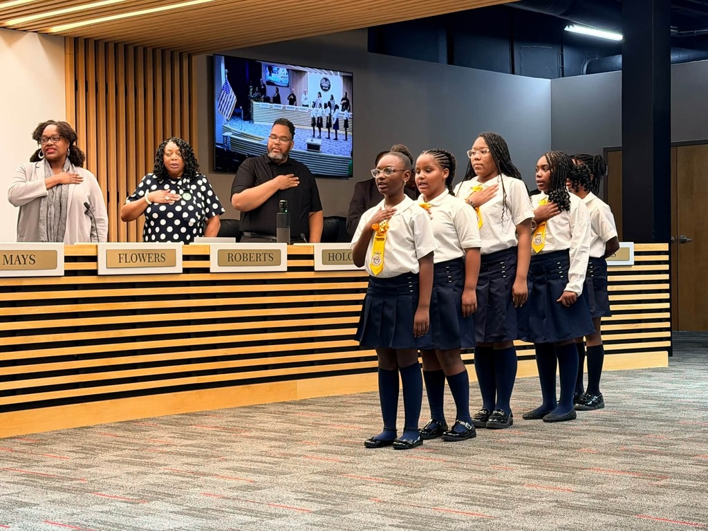 Seven young girls in school uniforms stand before a podium with a monitor in the background.