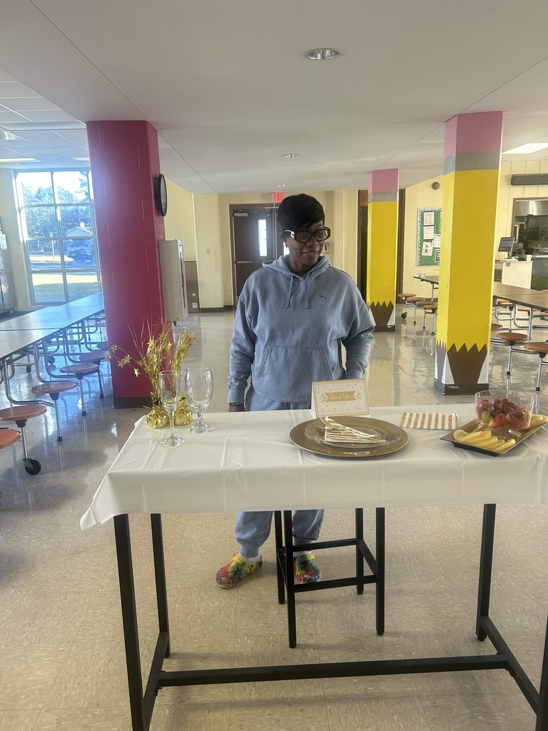 A person stands behind a white table with a plate, flowers, and other items on it in a room.