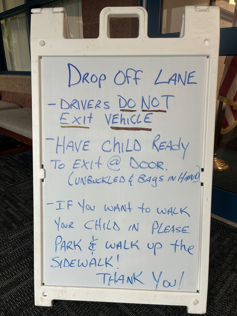 White sandwich board sign with blue handwritten text that reads: “Drop Off Lane. – Drivers DO NOT exit vehicle. – Have child ready to exit at door (unbuckled & bags in hand). – If you want to walk your child in, please park & walk up the sidewalk! Thank you.”