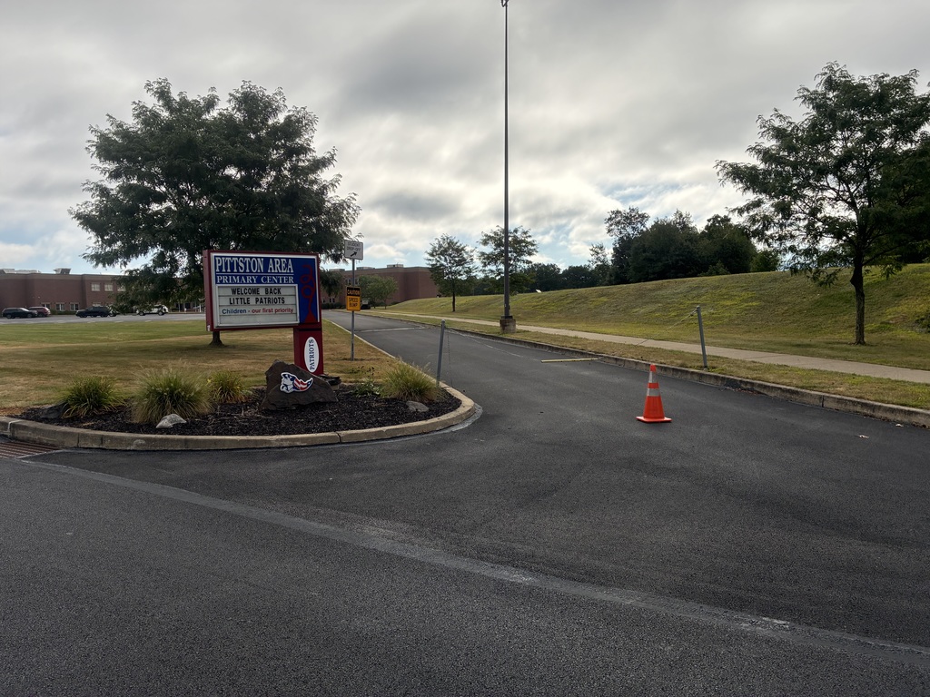 Entrance road to Pittston Area Primary Center. A red and blue school sign reads: “Pittston Area Primary Center – Welcome Back Little Patriots – Children, our first priority.” An orange cone is placed in the driveway marking the entrance lane.