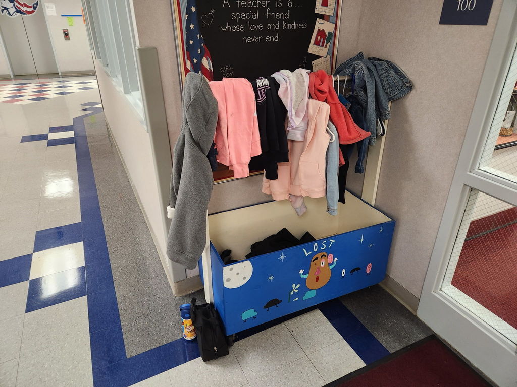The photo shows a school hallway with a Lost and Found area. The Lost and Found is a blue wooden box decorated with drawings, including a face, flowers, and stars, with the word "LOST" written across the front. Several jackets and sweaters are draped over the top and hanging on hooks, including pink, gray, and denim items. Inside the box, there are more clothing items. On the floor next to the box are a water bottle and a small black bag. The hallway floor has gray tiles with blue geometric patterns, and a classroom door with a window is visible to the right.