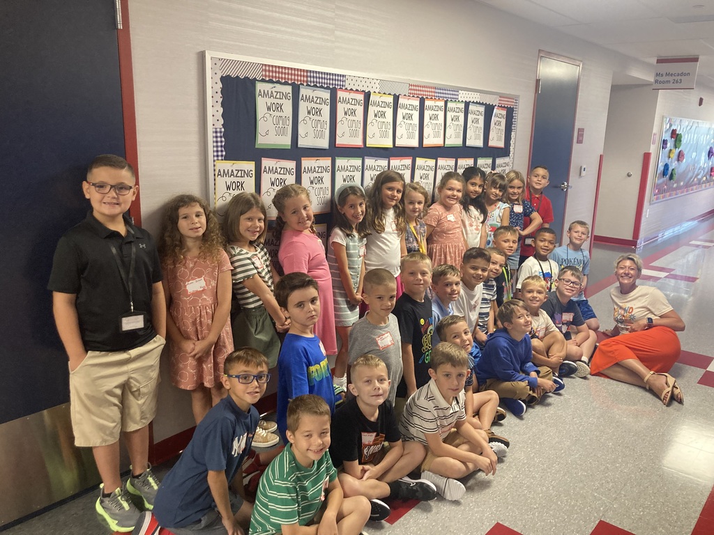 A large group of elementary school students poses together in a school hallway. Some students are standing, while others are kneeling or sitting on the floor. A teacher in an orange skirt lies on the floor smiling with the group. Behind them, a bulletin board displays the phrase “Amazing Work Coming Soon” in repeated colorful posters.