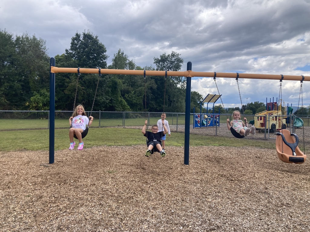 Students are swinging on a playground swing set with additional playground equipment in the background frwaturing a bus climber a blue sound wall and a small jungle gym.