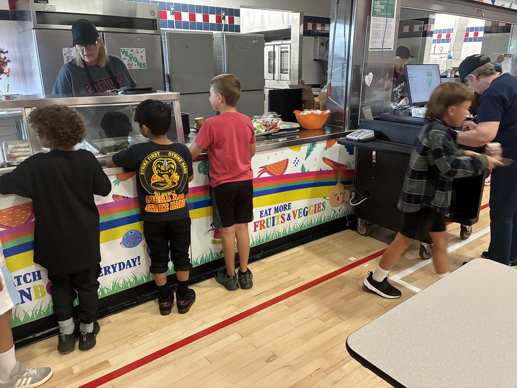 A group of young children stand in line at a school cafeteria counter, choosing their lunches. A cafeteria worker wearing a black cap and glasses serves food behind the counter. The counter is decorated with colorful signs promoting fruits and vegetables. Another staff member helps a child at a checkout station on the right.