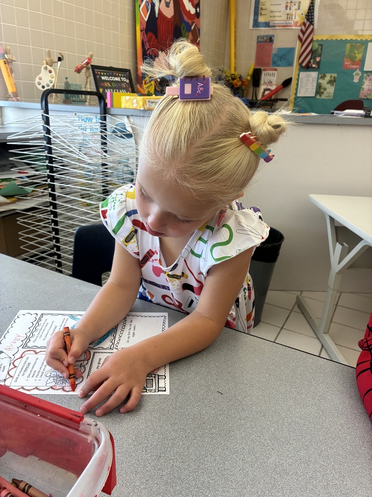 A girl colors on an activity sheet that introduces art class procedures. The activity sheet features a mona lisa. The girl is seated at a gray table in an art room using an orange crayon to color.
