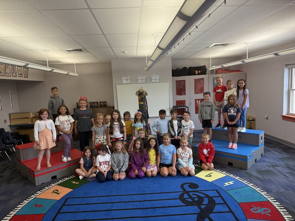 A classroom of young students poses together in a music room. Some children stand on red and blue risers, while others sit on a circular rug decorated with piano keys and musical notes. A teacher or adult is not visible in the photo, and the students are smiling at the camera.