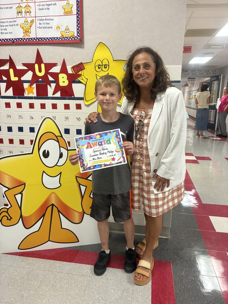 Spencer R. holding a colorful certificate of award stands next to a smiling Mrs. Rava in a school hallway. Behind them is a bulletin board decorated with large yellow cartoon stars, red stars, and motivational classroom posters.