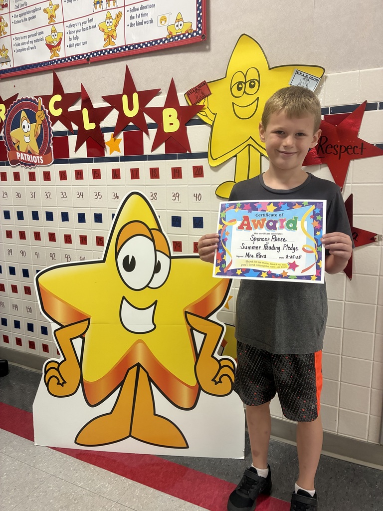 Spencer R. stands in a school hallway, smiling and holding a colorful certificate of award. Behind him is a bulletin board decorated with large yellow cartoon stars and red stars.