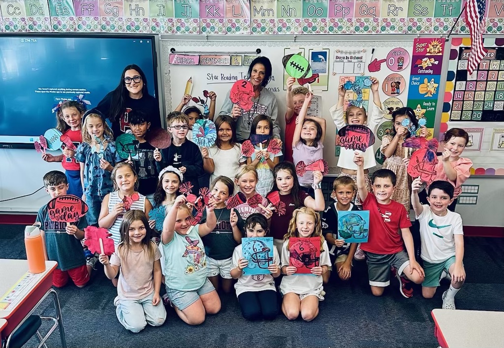 A classroom of smiling elementary students poses with two teachers. The children hold up colorful handmade football-themed signs and pom-poms in red and blue. The whiteboard behind them reads “Friday, September 5, 2025” with a school assignment listed.