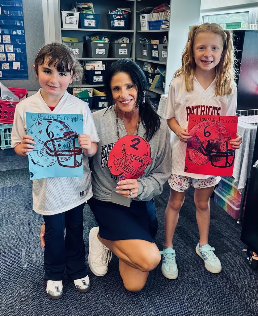 Lori Baumes kneels between two smiling students, Angelina F and Willow C. all showing off decorated football helmet posters in red and blue with encouraging messages for the team.