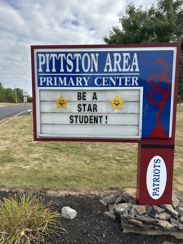 A rectangular outdoor school sign with a blue and white header reads “PITTSTON AREA PRIMARY CENTER” in bold white letters on a blue background. Below that, a changeable letter board displays the message “BE A STAR STUDENT!” with two yellow smiling star graphics on either side of the words. The bottom of the sign’s post is red with a white oval containing the word “PATRIOTS” in blue capital letters. The sign is set on a landscaped area with mulch, rocks, and some plants. A road and grassy area with trees are visible in the background under a cloudy sky.