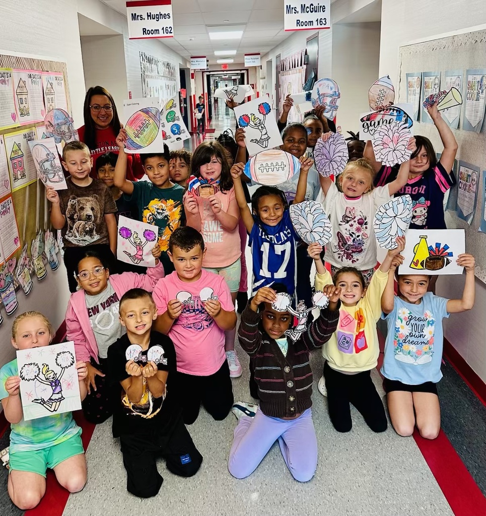 A group of about 20 second-grade students and their teacher are standing and kneeling in a brightly lit school hallway. They are smiling and proudly holding up handmade, colorful drawings and signs featuring footballs, pom-poms, megaphones, and cheerleader designs. Above them, classroom signs read “Mrs. Hughes Room 162” and “Mrs. McGuire Room 163.” The students appear happy and excited, showing their support for the Patriot football team.