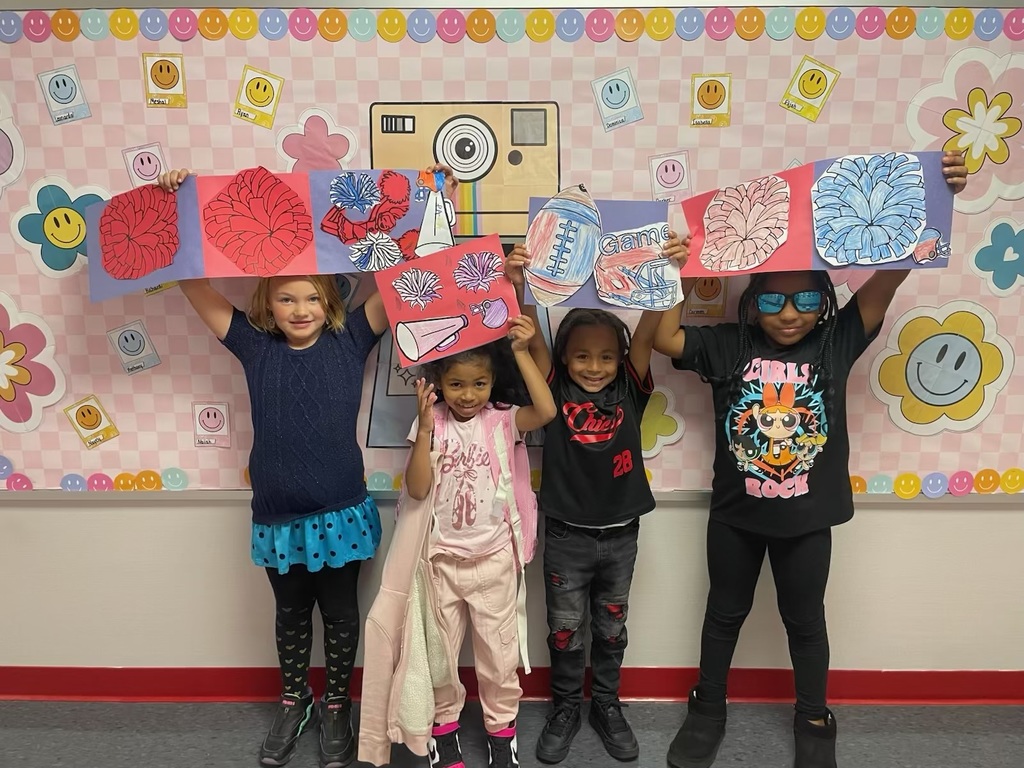 Four students standing in a group in front of a pink smiley and floral bulletin board. They are holding up red and blue construction paper signs decorated with pom poms, football helmets and fottballs.