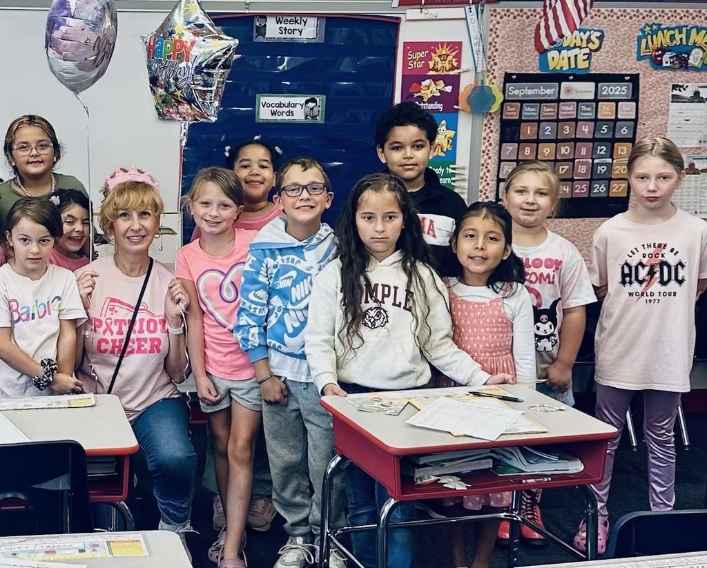 A group of students posing in their classroom with their teacher Mrs. Chesneak. Mrs. Chesneak is seated in the middle of the group of students with a pink birthday crown and two birthday balloons.