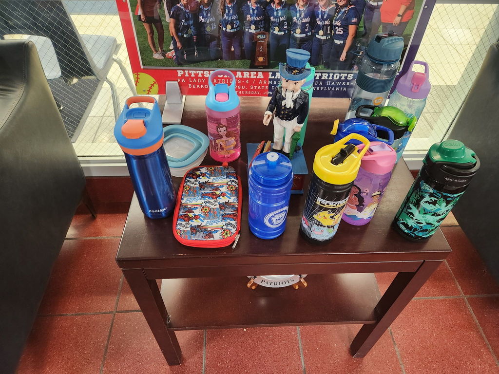 Brown wooden table in a school lobby with a red tile floor. The table has several children’s water bottles and a patriot sculpture.