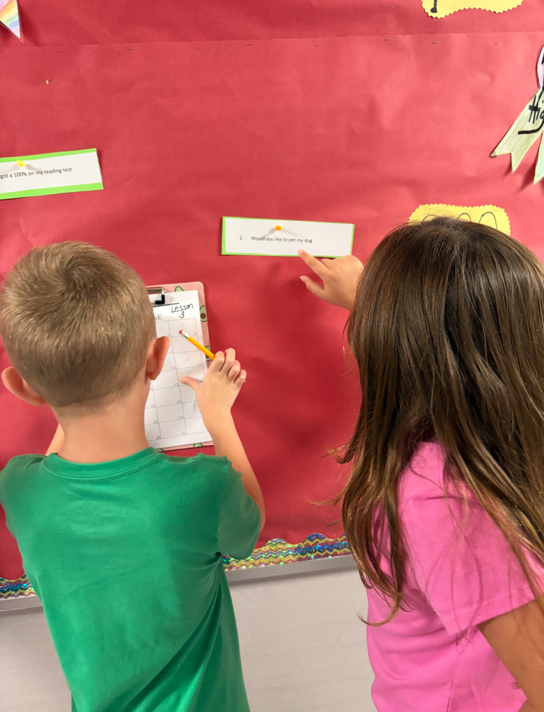 Students stand side by side at a brightly decorated bulletin board. One is pointing at a sentence card on the wall while the other holds a clipboard labeled ‘Footloose’ with numbered spaces for answers. Both are actively participating in a movement-based learning activity, reading and recording responses together.