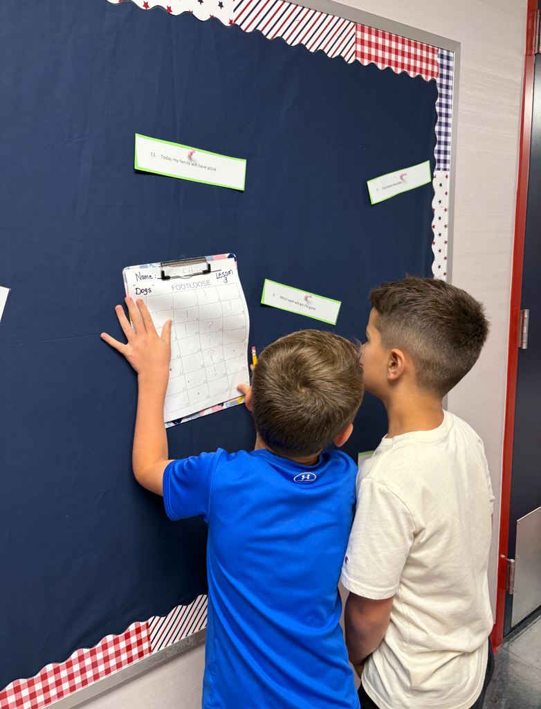 Students stand side by side at a brightly decorated bulletin board. One is pointing at a sentence card on the wall while the other holds a clipboard labeled ‘Footloose’ with numbered spaces for answers. Both are actively participating in a movement-based learning activity, reading and recording responses together.