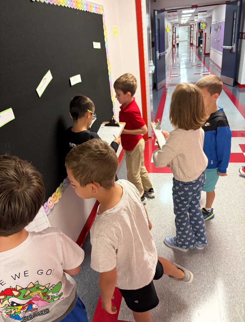 Students stand side by side at a brightly decorated bulletin board. One is pointing at a sentence card on the wall while the other holds a clipboard labeled ‘Footloose’ with numbered spaces for answers. Both are actively participating in a movement-based learning activity, reading and recording responses together.