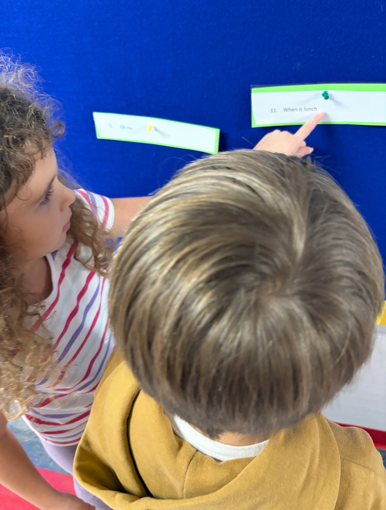 Students stand side by side at a brightly decorated bulletin board. One is pointing at a sentence card on the wall while the other holds a clipboard labeled ‘Footloose’ with numbered spaces for answers. Both are actively participating in a movement-based learning activity, reading and recording responses together.
