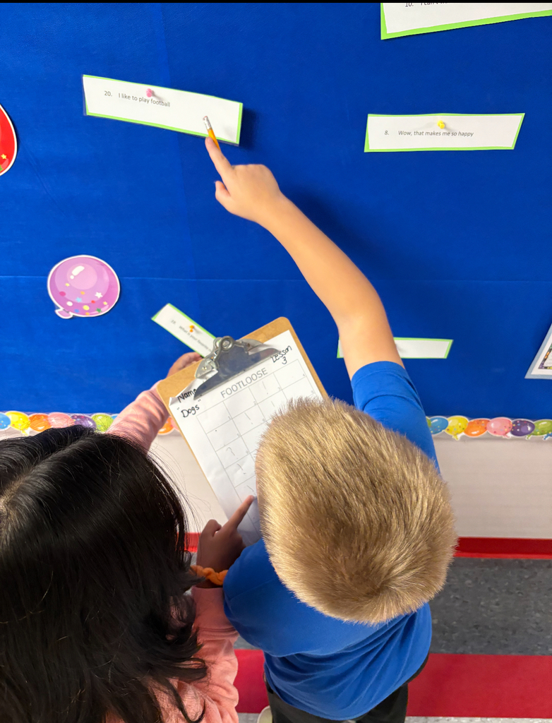Students stand side by side at a brightly decorated bulletin board. One is pointing at a sentence card on the wall while the other holds a clipboard labeled ‘Footloose’ with numbered spaces for answers. Both are actively participating in a movement-based learning activity, reading and recording responses together.
