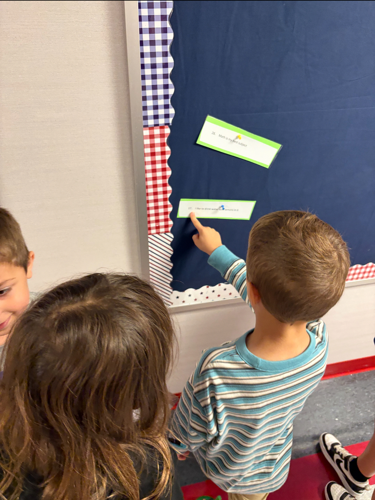 Students stand side by side at a brightly decorated bulletin board. One is pointing at a sentence card on the wall while the other holds a clipboard labeled ‘Footloose’ with numbered spaces for answers. Both are actively participating in a movement-based learning activity, reading and recording responses together.