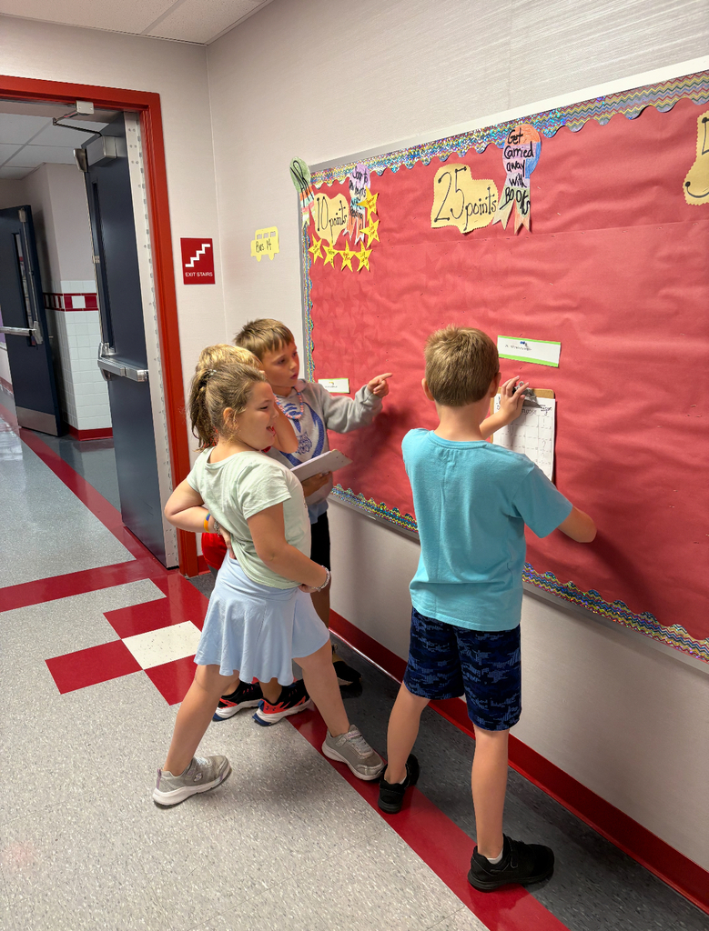 Students stand side by side at a brightly decorated bulletin board. One is pointing at a sentence card on the wall while the other holds a clipboard labeled ‘Footloose’ with numbered spaces for answers. Both are actively participating in a movement-based learning activity, reading and recording responses together.