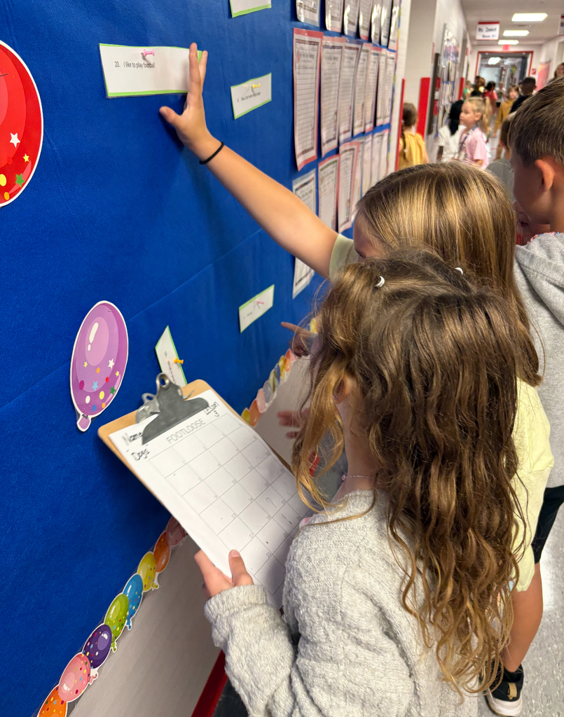 Students stand side by side at a brightly decorated bulletin board. One is pointing at a sentence card on the wall while the other holds a clipboard labeled ‘Footloose’ with numbered spaces for answers. Both are actively participating in a movement-based learning activity, reading and recording responses together.