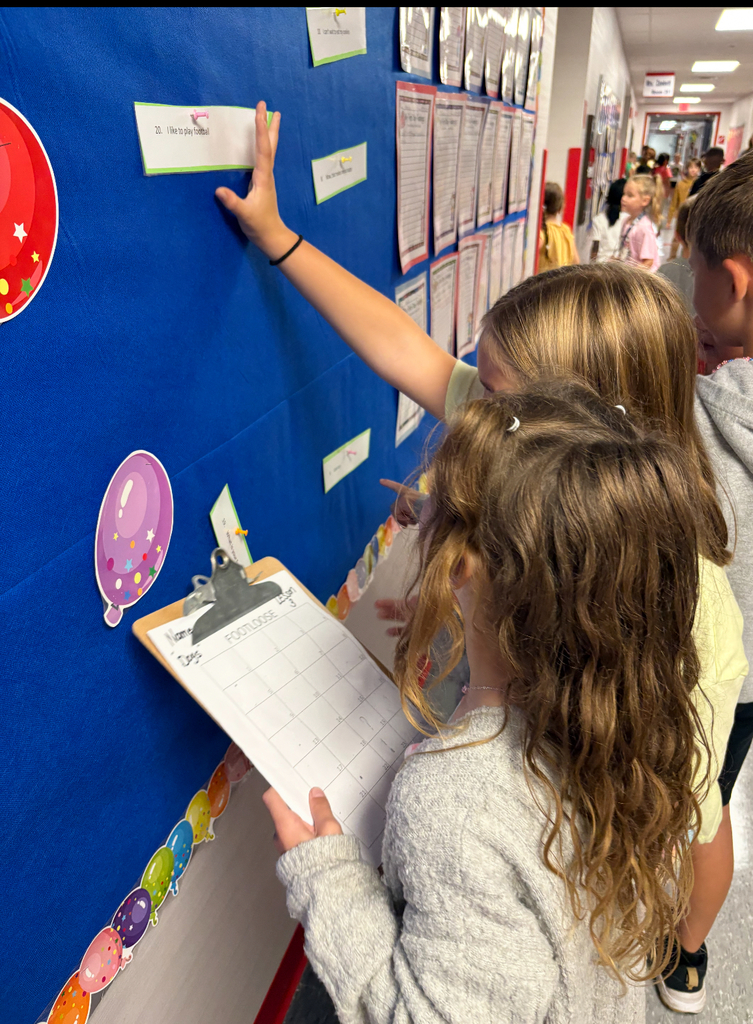 Students stand side by side at a brightly decorated bulletin board. One is pointing at a sentence card on the wall while the other holds a clipboard labeled ‘Footloose’ with numbered spaces for answers. Both are actively participating in a movement-based learning activity, reading and recording responses together.